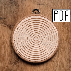 以下が含まれることがあります： ベージュの背景に、金色のアクセントがついた白いビーズの同心円が描かれた円形の刺繍枠。枠は木製の表面の上に置かれています。PDF