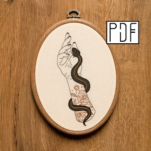 Può includere: Un telaio da ricamo ovale contenente un pannello in tessuto con un serpente nero e arancione che si avvolge attorno a una mano e un avambraccio. L'avambraccio presenta un ricamo floreale a linee. Il testo PDF è visibile in alto a destra.