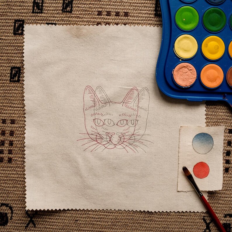 May include: A white fabric square with a red line drawing of a cat with four eyes. The cat is looking directly at the viewer. The fabric is on a brown woven surface. There is a blue and white watercolor palette and a paintbrush on the surface.