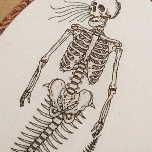 Puede incluir: Un bordado detallado sobre tela color hueso que muestra un esqueleto marr&oacute;n oscuro con una cola de sirena. El esqueleto se representa desde el torso hacia arriba, con l&iacute;neas fluidas que salen de la boca, dentro de un bastidor de madera.