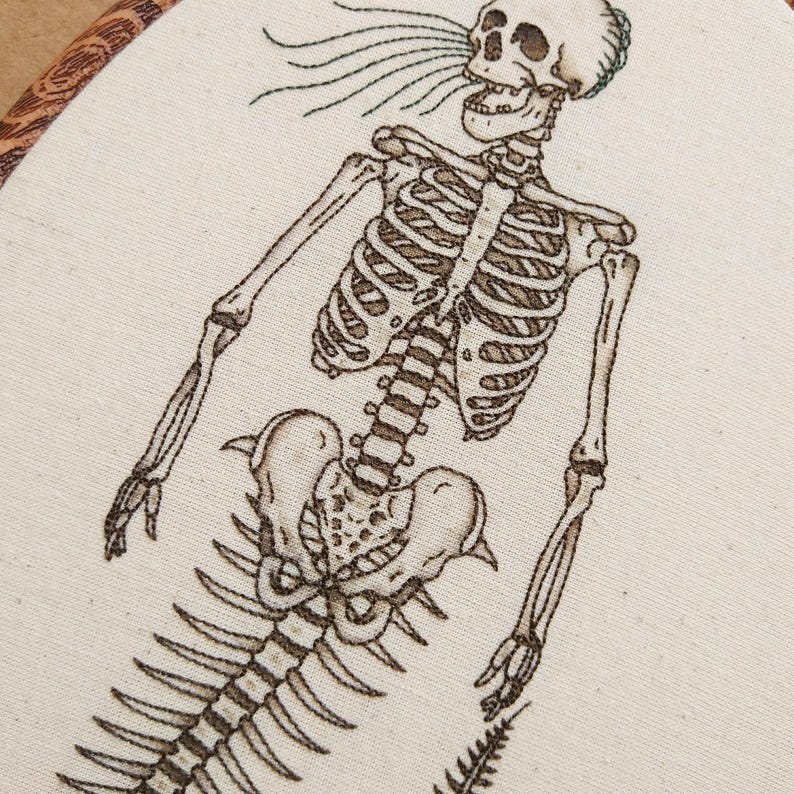 Puede incluir: Un bordado detallado sobre tela color hueso que muestra un esqueleto marr&oacute;n oscuro con una cola de sirena. El esqueleto se representa desde el torso hacia arriba, con l&iacute;neas fluidas que salen de la boca, dentro de un bastidor de madera.