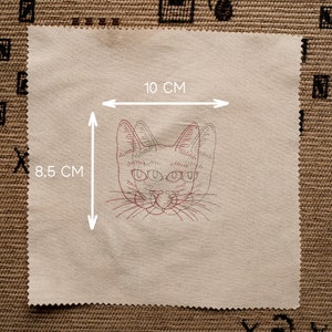 May include: A beige fabric patch with a red line drawing of a cat's face with three eyes. The fabric patch measures 8.5 cm by 10 cm.