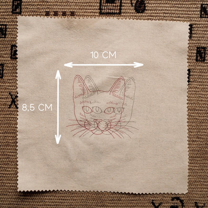 May include: A beige fabric patch with a red line drawing of a cat's face with three eyes. The fabric patch measures 8.5 cm by 10 cm.
