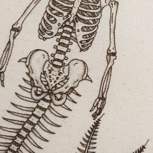 Puede incluir: Un dise&ntilde;o de esqueleto bordado en marr&oacute;n sobre un fondo de tela color crema. El bordado muestra la parte superior del torso, la caja tor&aacute;cica, la columna, la pelvis y los huesos de los brazos de un esqueleto humano, con un helecho abajo.