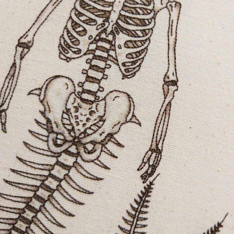 Puede incluir: Un dise&ntilde;o de esqueleto bordado en marr&oacute;n sobre un fondo de tela color crema. El bordado muestra la parte superior del torso, la caja tor&aacute;cica, la columna, la pelvis y los huesos de los brazos de un esqueleto humano, con un helecho abajo.