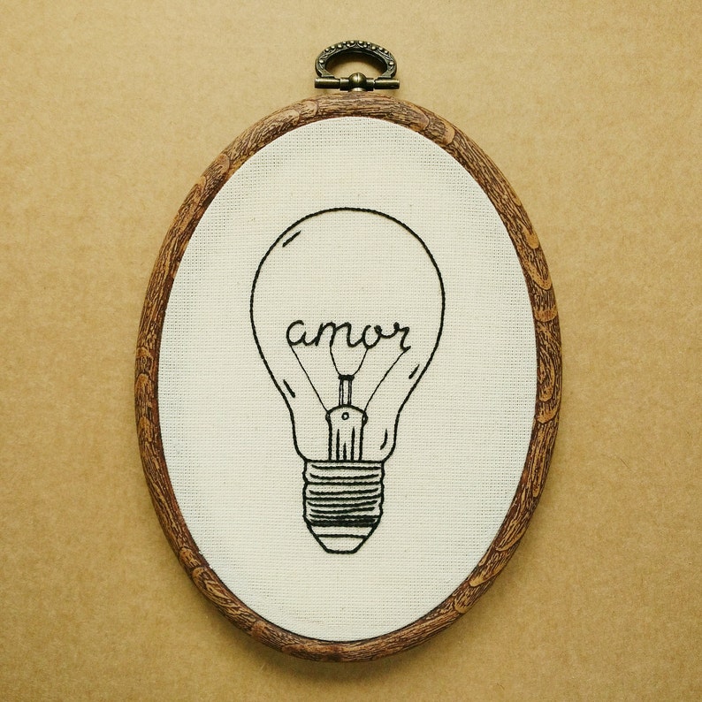 May include: Oval embroidery hoop featuring a lightbulb design with the word "amor" stitched in black thread on white fabric. The wooden hoop has a metal loop for hanging.