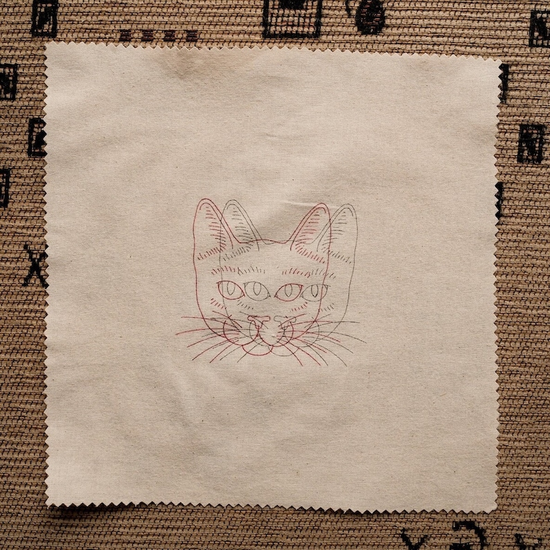May include: A beige fabric square with a red line drawing of a cat's face with four eyes. The cat's face is centered on the fabric square.