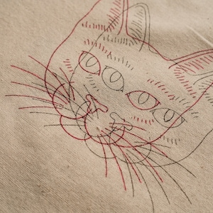 May include: A close-up of a red and black line drawing of a cat's face on a beige fabric background.