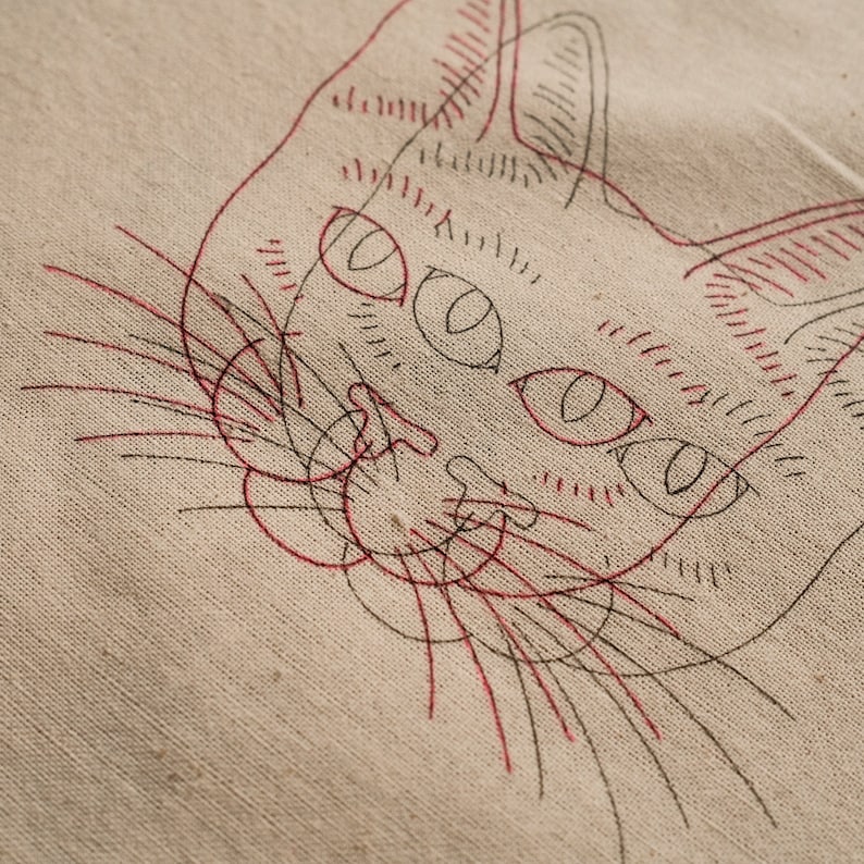 May include: A close-up of a red and black line drawing of a cat's face on a beige fabric background.