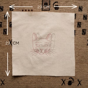 May include: A beige fabric square with a red and black embroidered cat face with three eyes. The fabric is 23 inches by 23 inches.