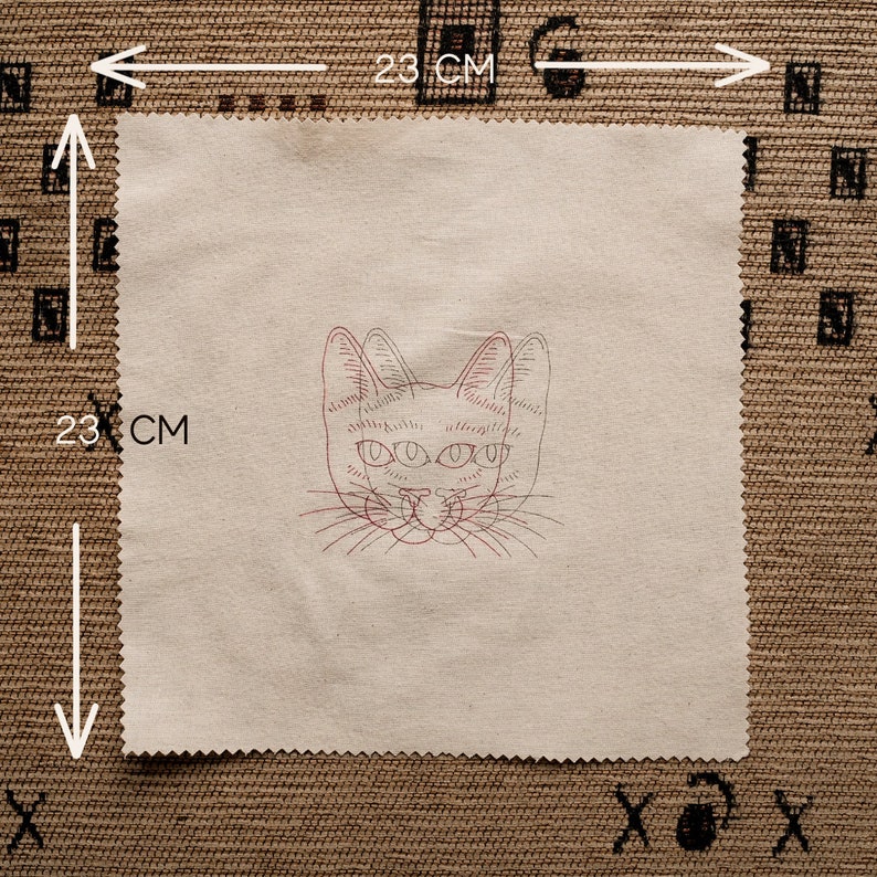 May include: A beige fabric square with a red and black embroidered cat face with three eyes. The fabric is 23 inches by 23 inches.
