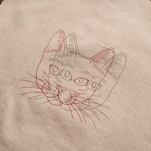 May include: A beige fabric with a red and black line drawing of a cat's face. The cat has large eyes and a long nose.