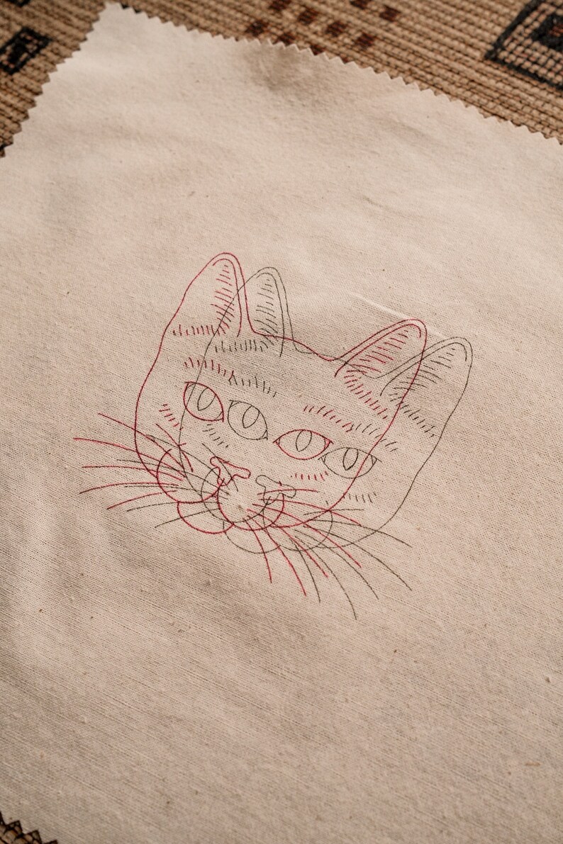 May include: A beige fabric with a red and black line drawing of a cat's face. The cat has large eyes and a long nose.