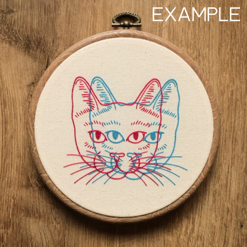 May include: Embroidered cat face with red and blue thread in a wooden embroidery hoop. The cat has four eyes and a 3D effect.