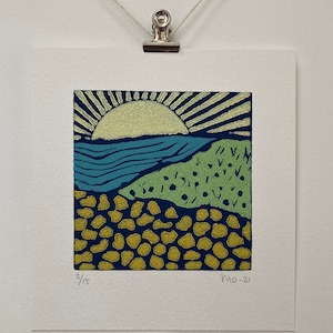 May include: A colorful linocut print of a sunrise over a beach scene. The print features a yellow sun with rays, a blue ocean, green grass, and a yellow and blue patterned beach. The print is numbered 3/15 and dated MO-21.