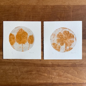 May include: Two square white paper prints featuring circular orange botanical leaf and flower imprints. The left print shows three leaves, and the right print shows a flower cluster. Each print includes a handwritten signature and date.