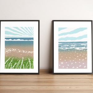 May include: Two framed prints of seascapes. The print on the left shows a beach with green grass and a blue sky with sun rays. The print on the right shows a beach with white sand and a blue sky with clouds.