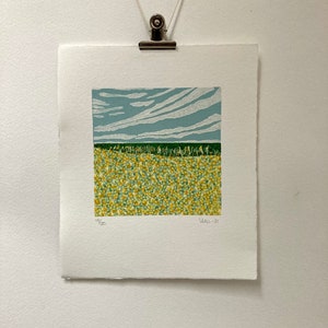 May include: A print of a landscape with a blue sky and white clouds, a green field, and a yellow field with small dots. The print is numbered 19/20 and dated 2021.