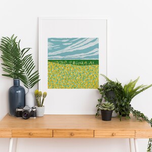May include: A framed print of a landscape with a field of yellow flowers, a green field, and a blue sky with white clouds. The print is on a wooden desk with a vase of green leaves, a camera, and two potted plants.