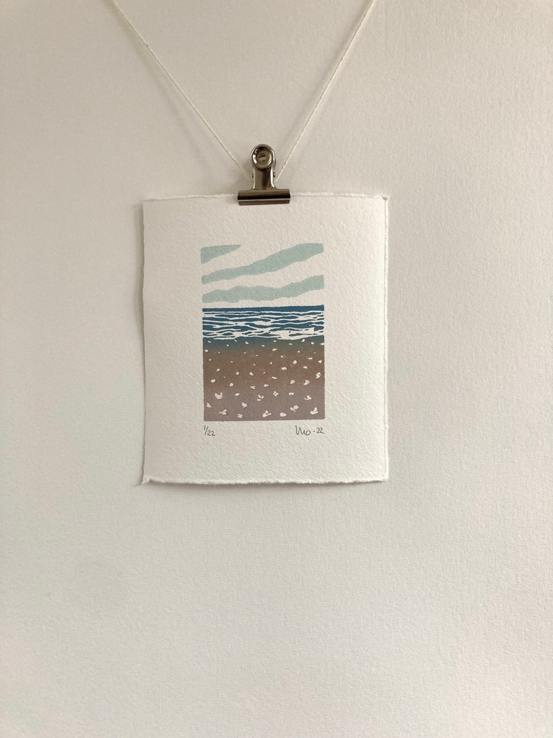 May include: A minimalist print of a seascape with a blue ocean, brown sand, and white clouds against a light blue sky. The print is signed and dated "1/22  Nov-22".
