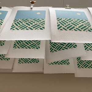 May include: A stack of white paper prints with a green and white geometric pattern and a blue sky with a white circle. The prints are hanging from a wall with clips.