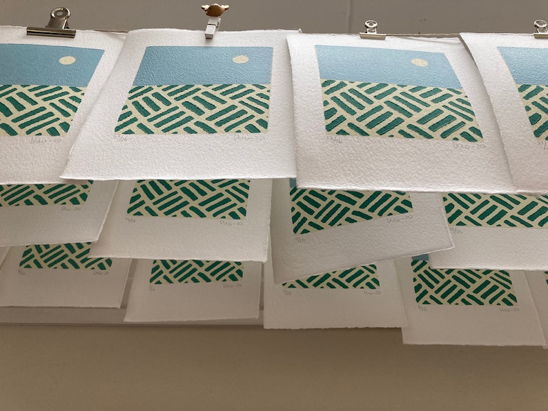 May include: A stack of white paper prints with a green and white geometric pattern and a blue sky with a white circle. The prints are hanging from a wall with clips.