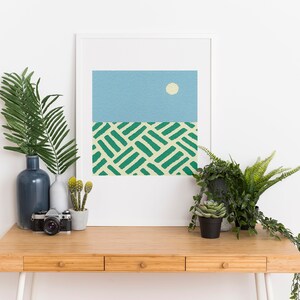 May include: A framed abstract print with a blue sky and a white sun, above a green and yellow patterned field. The print is on a wooden desk with a vase of green leaves, a camera, and potted plants.