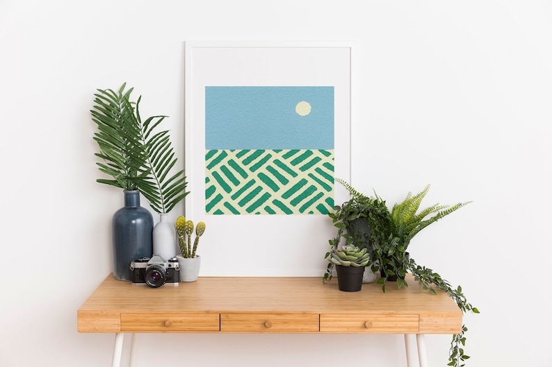 May include: A framed abstract print with a blue sky and a white sun, above a green and yellow patterned field. The print is on a wooden desk with a vase of green leaves, a camera, and potted plants.