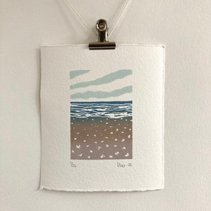 May include: A blue and white abstract print of a seascape with a brown sandy beach. The print is numbered 1/22 and dated Nov. 22.