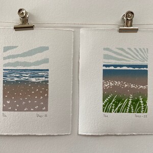 May include: Two framed prints of seascapes with a blue sky, ocean, and beach. The print on the left has a simple sky with clouds, while the print on the right has a sunburst sky. Both prints have the text "1/22" and "Wo-22" in the bottom right corner.