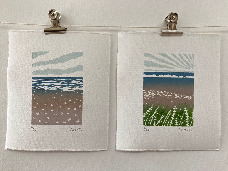 May include: Two framed prints of seascapes with a blue sky, ocean, and beach. The print on the left has a simple sky with clouds, while the print on the right has a sunburst sky. Both prints have the text "1/22" and "Wo-22" in the bottom right corner.