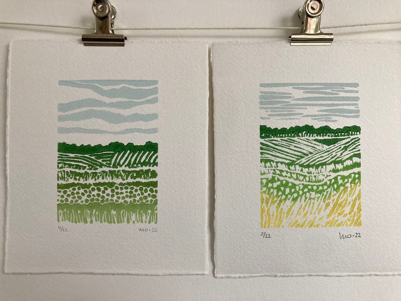 May include: Two framed prints of green and yellow landscape scenes with a blue sky. The prints are signed and dated "4/22" and "11.10.22".