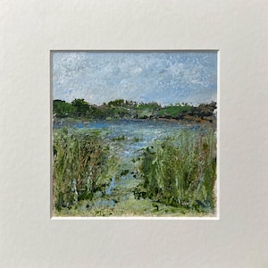 May include: An original oil painting of a landscape scene. The artwork depicts a body of water with green reeds in the foreground, a line of green trees, and a cloudy blue sky. The painting is framed in a white mount.