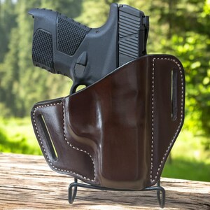 May include: A black handgun is partially inserted into a brown leather holster. The holster is supported by a black metal stand. The leather holster has white stitching around the edges. The background is a blurred view of a forest.