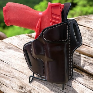 May include: A red handgun is partially inserted into a black leather holster. The holster has white stitching around the edges. The handgun and holster are resting on a wooden surface, with a blurred green background.