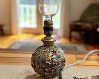 May include: A gold-colored lamp base decorated with rhinestones, beads, and other embellishments. The lamp has a clear glass globe shade and a gold-colored cord.