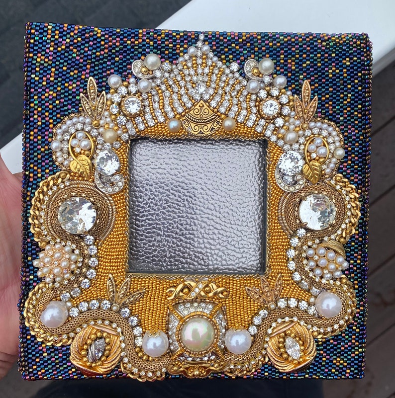 May include: A gold picture frame with a beaded design. The frame is decorated with pearls, rhinestones, and gold beads. The frame is on a dark blue background.