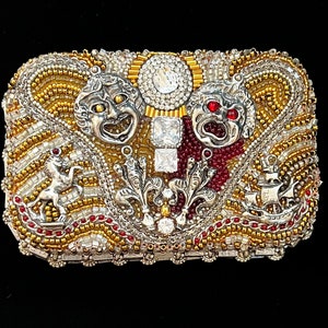 May include: A silver and gold beaded box with a design of two theatrical masks, a horse, and a ship. The masks are silver with red and white accents. The box is decorated with rhinestones.