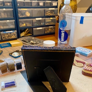 May include: A black picture frame with a beaded border sits on a table with various craft supplies, including small containers of beads, a clear water bottle labeled "smartwater", and a small decorative box.