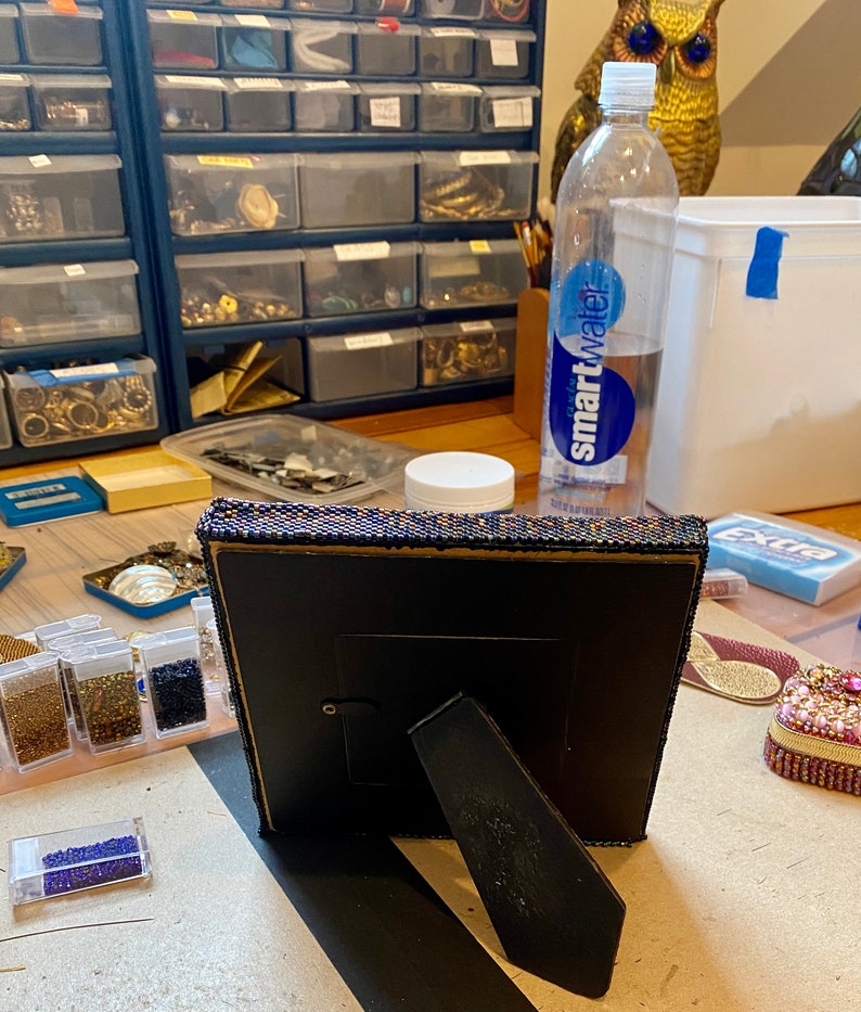 May include: A black picture frame with a beaded border sits on a table with various craft supplies, including small containers of beads, a clear water bottle labeled "smartwater", and a small decorative box.