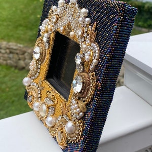 May include: A small, dark blue picture frame with a gold and white beaded design. The frame is decorated with pearls, rhinestones, and gold accents.