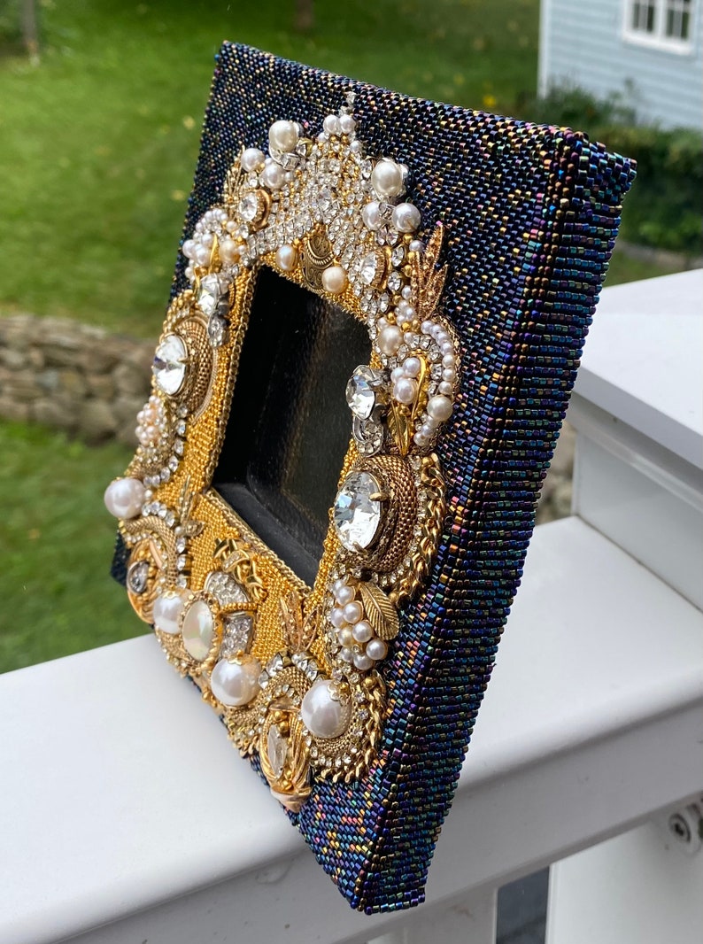 May include: A small, dark blue picture frame with a gold and white beaded design. The frame is decorated with pearls, rhinestones, and gold accents.