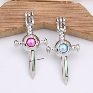 May include: Two silver-colored sword pendants with cross-shaped designs. One pendant features a pink pearl, the other a blue pearl. The sword blades have a green stripe. The Etsy shop name "LDjewelrycharm.Etsy" is visible.