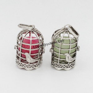 May include: Two silver-colored birdcage pendants, each with a small bird perched on the front. One cage holds a pink sphere, the other a green sphere. The cages have a decorative base and a loop for hanging.