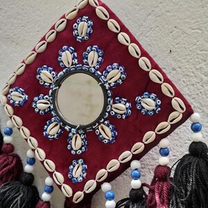 May include: A burgundy square wall hanging with a round mirror in the center. The mirror is surrounded by white and blue seashells. The hanging has tassels in shades of burgundy, black, and blue.