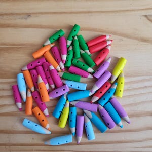 May include: A collection of colorful pencil-shaped erasers in various colors, including green, pink, orange, blue, yellow, and purple. The erasers are scattered on a light brown wooden surface. Each eraser has a hole in the middle.