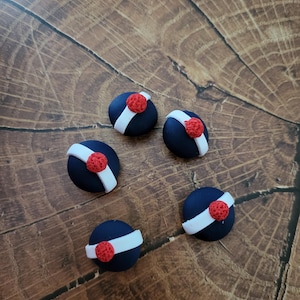 Può includere: Cinque bottoni decorativi blu navy con una striscia bianca e una pallina rossa. Sono disposti su una superficie di legno con un motivo a venature naturali. I bottoni sono probabilmente per progetti di artigianato o cucito.