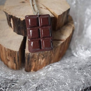 Peut inclure: Un collier avec un pendentif en forme de barre de chocolat marron sur une chaîne argentée. Le pendentif a la forme d'une barre de chocolat avec des carrés et repose sur une surface en bois.