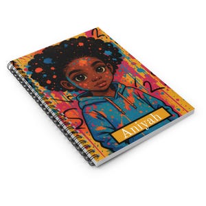 May include: Spiral-bound notebook featuring a colorful illustration of a young person with dark skin wearing a blue hoodie. The notebook cover has a vibrant, abstract background with the name "Aniyah" on a yellow rectangle.