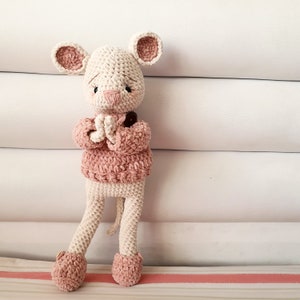 May include: A crocheted mouse toy wearing a pink sweater. The mouse has a white body and pink ears and nose. The toy is standing on a white and pink striped surface.